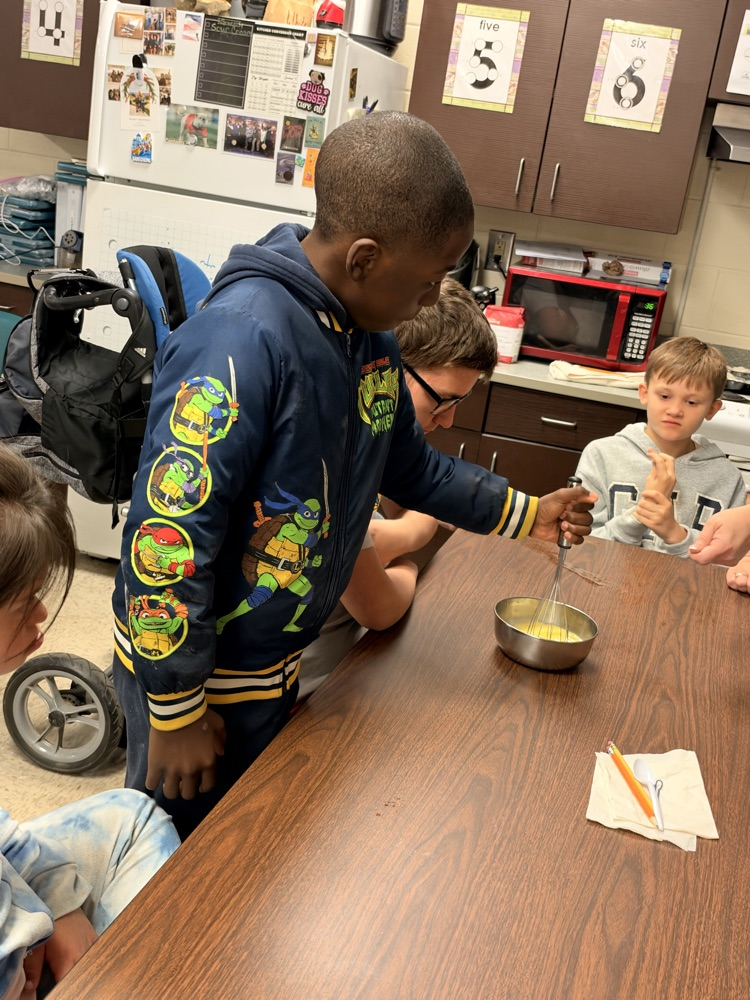 A boy with a blue hoodie stirs a bowl of food while others sit around a table.