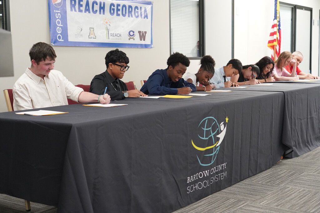 Students sit at a table with black tablecloths, writing on paper. A flag is visible in the background.