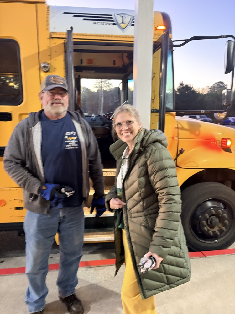 Two adults, one in a blue glove, stand before a yellow school bus at night.