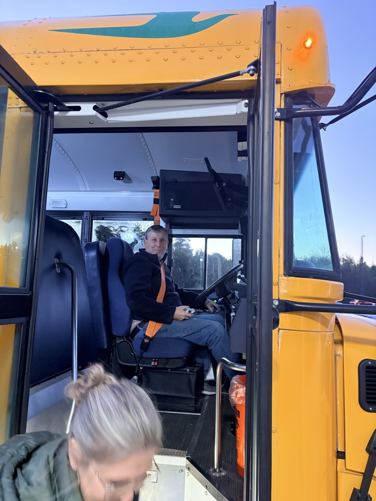 A woman with a ponytail views a school bus interior. A person in a jacket sits in the driver's seat.