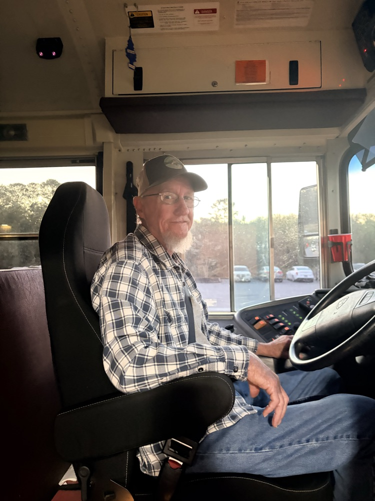 Man wearing a plaid shirt and cap sits in a bus driver's seat, steering wheel in hand.