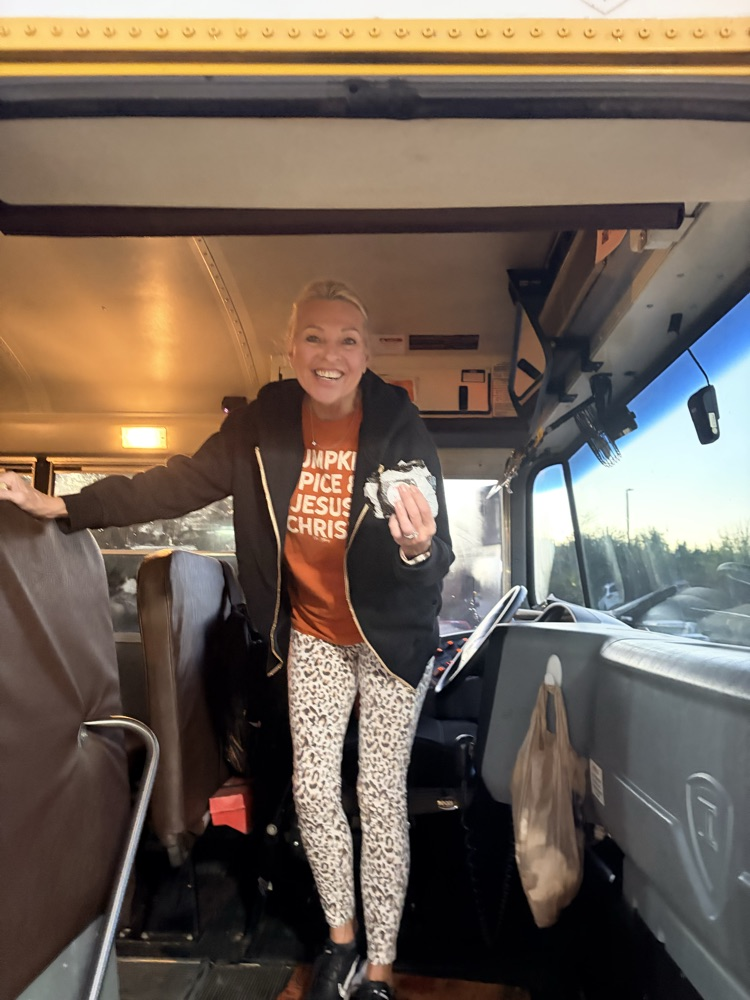 Woman inside school bus, smiling, holding food, standing at entrance with driver's seat visible.