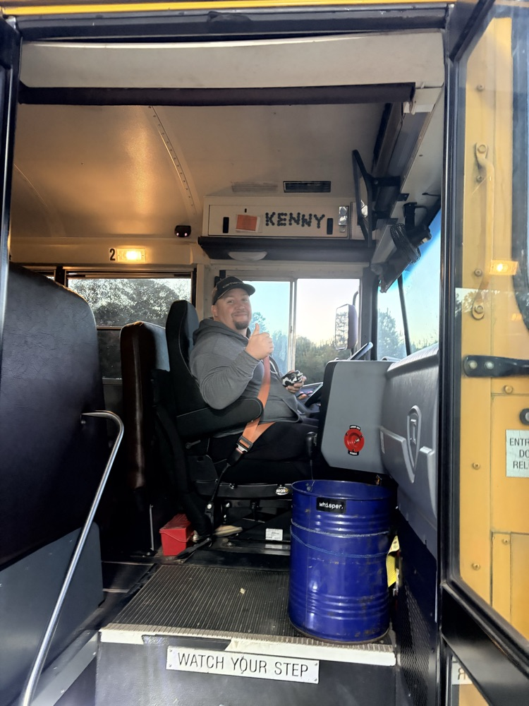 A man sits in the driver's seat of a yellow school bus with a blue barrel on the floor.