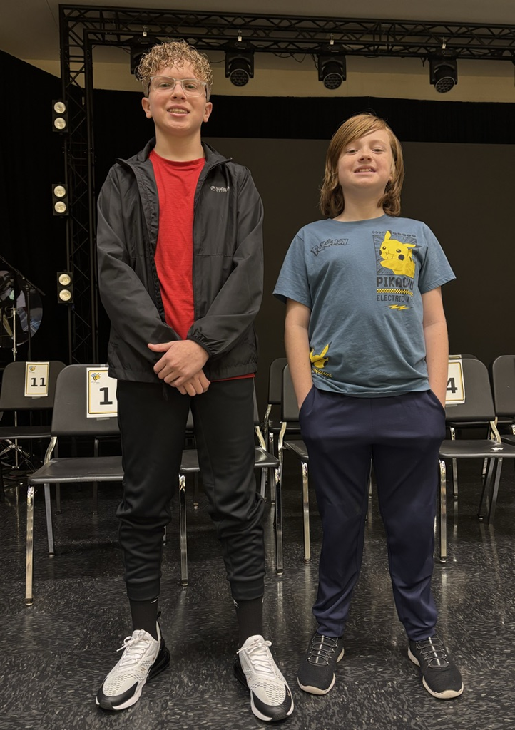 Two children, one in a red shirt and the other in a blue shirt, standing on a stage with chairs.