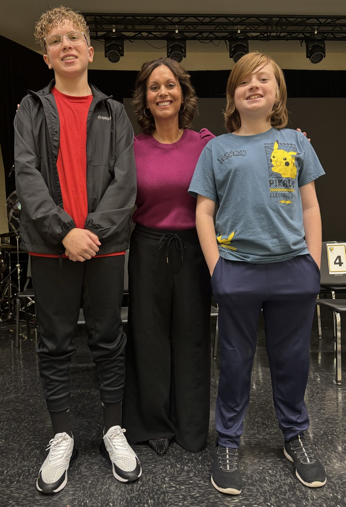 Three people standing on a stage, two boys and a woman. Behind them is a drum set and chairs.