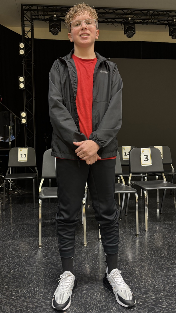 Boy in red shirt and black jacket standing in front of black backdrop, stage, chairs, drum set.