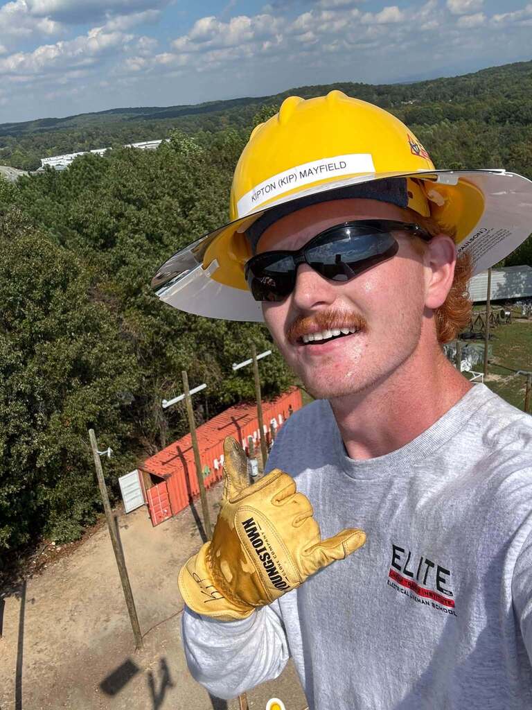 Man in yellow helmet, sunglasses, and yellow gloves pointing to camera, standing near utility poles.