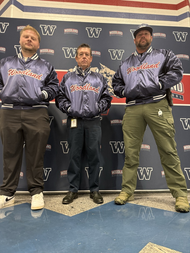 Three men wearing Woodland jackets and pants stand together in front of a backdrop with large letters.