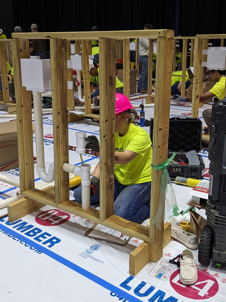A woman wearing yellow shirt and pink hat works on a wooden structure with pipework.