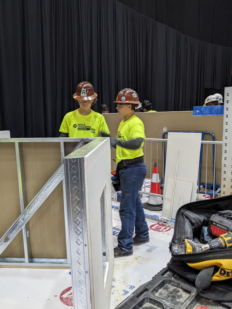 Two people in yellow hard hats and gloves work with tools and construction materials in an unfinished room.