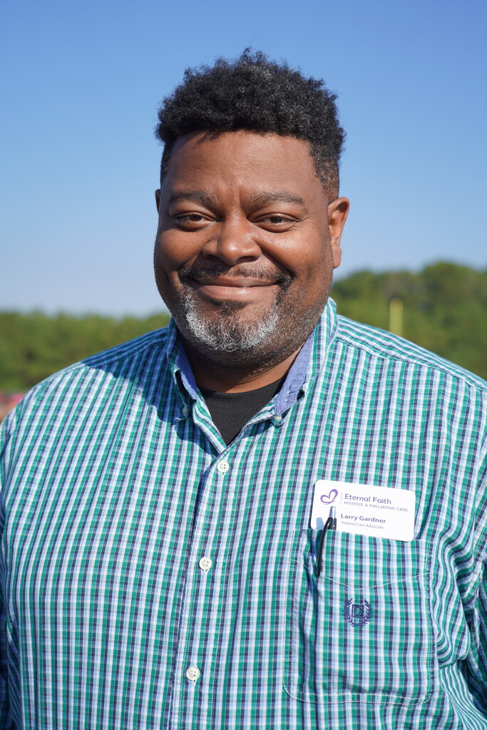 A man wearing a green and white checkered shirt smiles at the camera, with a white name tag in his chest pocket.