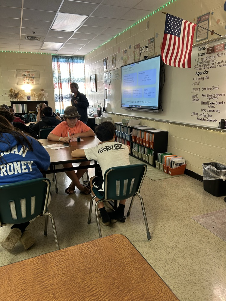 Students seated at desks inside a classroom, with a teacher standing at the front.