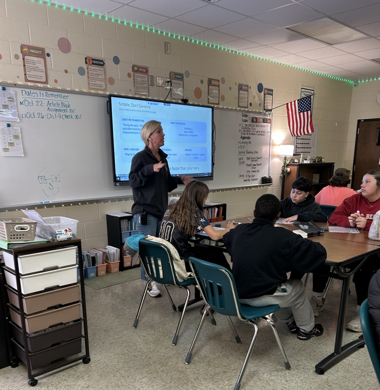 Classroom with students seated at tables, teacher stands before a projector screen, whiteboard with papers pinned.