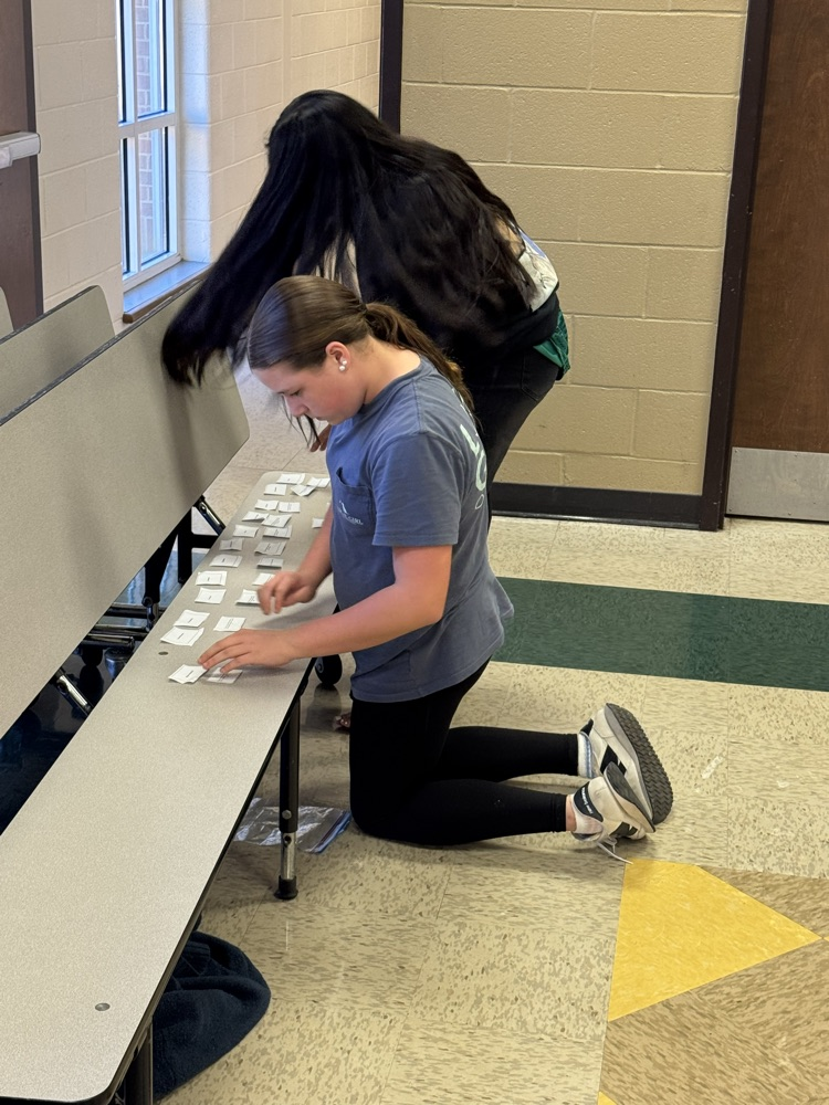 Two students interact with cards on a bench in a hallway with a brick wall.