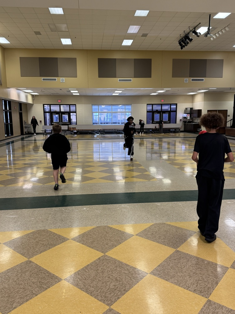 Indoor hallway with yellow and white checkered floor, people walking and standing, bright lighting, and stage at the end.