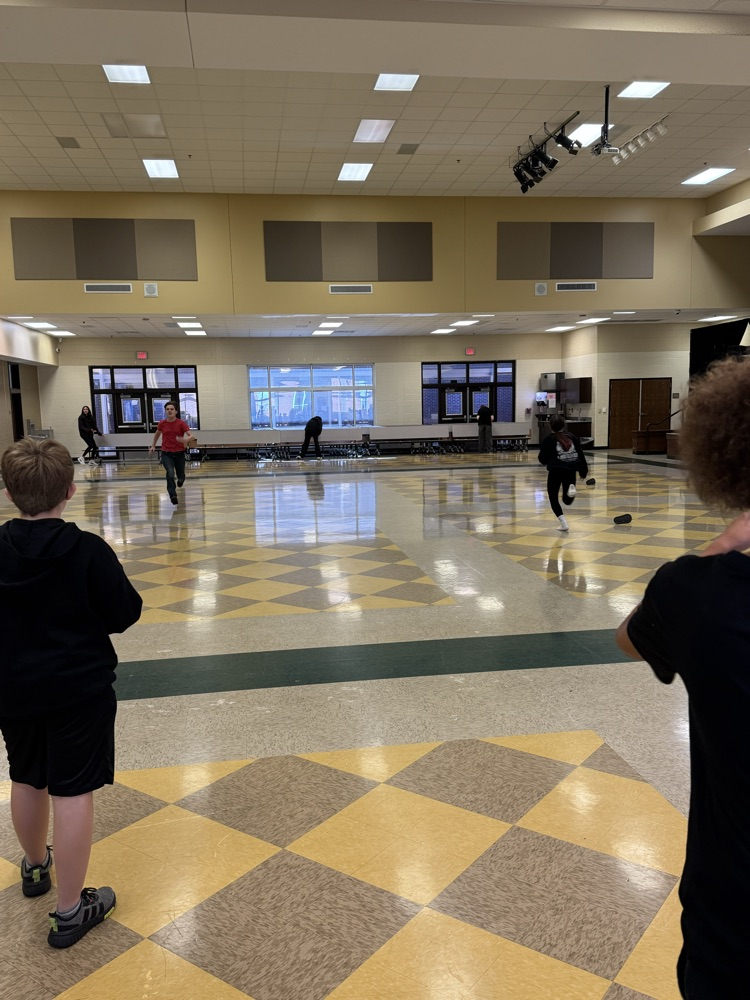 Indoor gymnasium with checkered floor. Several children in black shirts and sneakers are playing a game.