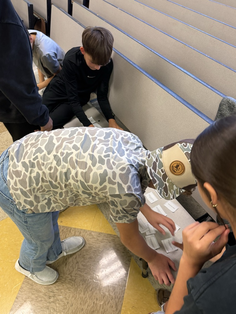 Students work on a floor project with one holding a board, others bending over, and one wearing a hat.