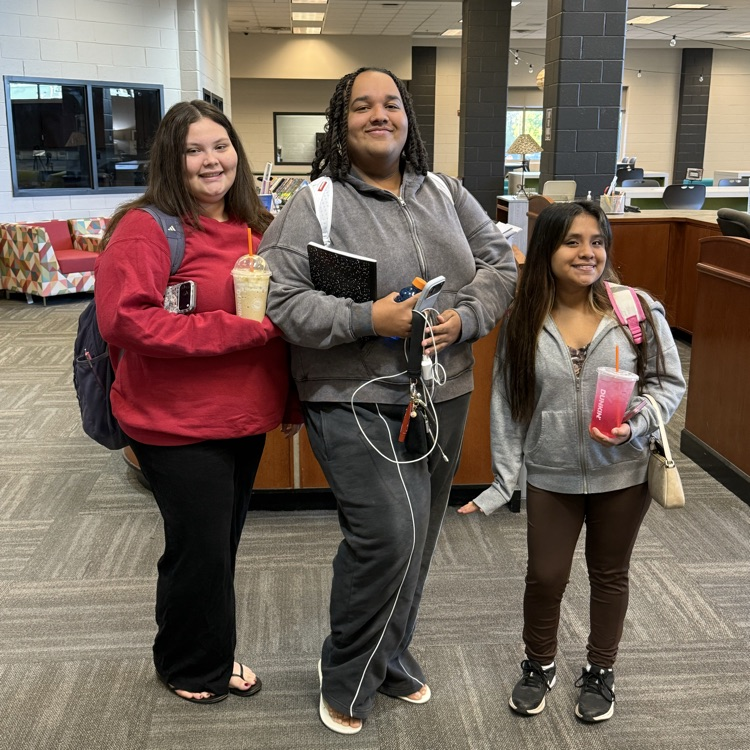Three students standing in a room with one holding a drink with a straw and a book.