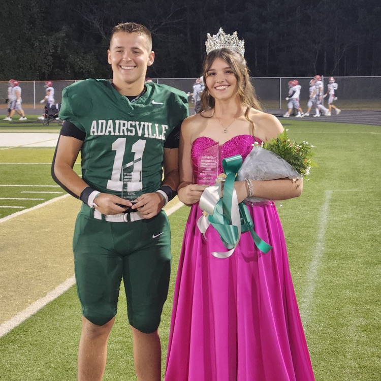 Teen boy and girl standing on a football field. Boy wearing green sports uniform. Girl wearing pink dress and holding bouquet.