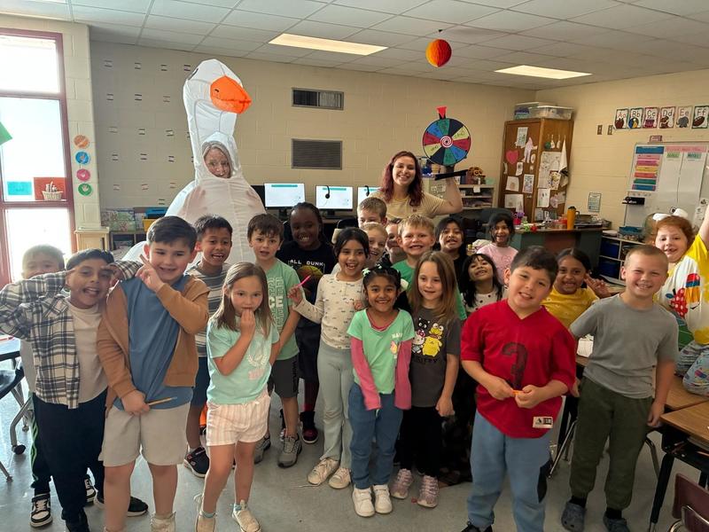 A group of elementary students stand together in a classroom with an adult wearing a large white goose costume. Another adult stands beside a colorful spinning wheel near computers and classroom materials.
