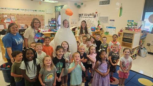 A group of elementary students and adults stand together in a decorated classroom, with one adult wearing a large white goose costume at the center. Students are arranged in rows in front of classroom shelves, posters, and learning materials.