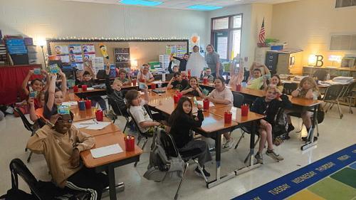 Students sit at desks arranged in rows inside a classroom with shelves, posters, and an exit sign on the back wall. Several students hold their arms up while looking toward the camera.