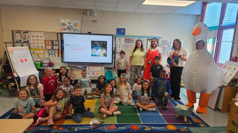 Elementary students sit and stand on a colorful classroom rug while adults stand behind them near a large interactive display screen. An adult in a white goose costume stands to the right near classroom posters and charts.