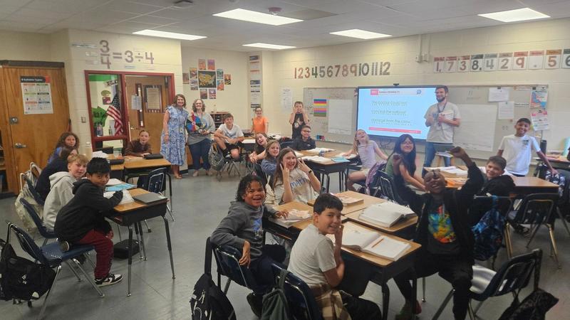 Students sit at desks arranged in groups inside a classroom decorated with hanging decorations and posters. Several adults stand at the front of the room near a presentation screen displaying colorful circular images.