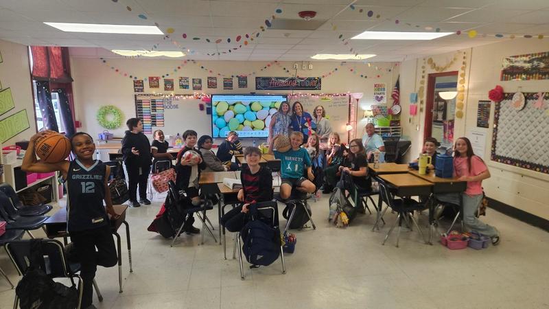 Students sit at individual desks facing the front of a classroom with number charts on the wall. Adults stand near a presentation screen and whiteboard at the front of the room.