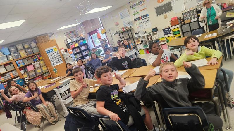 Students sit at grouped desks in a classroom with bulletin boards, storage bins, and classroom displays. An adult stands at the back of the room while students raise their hands.