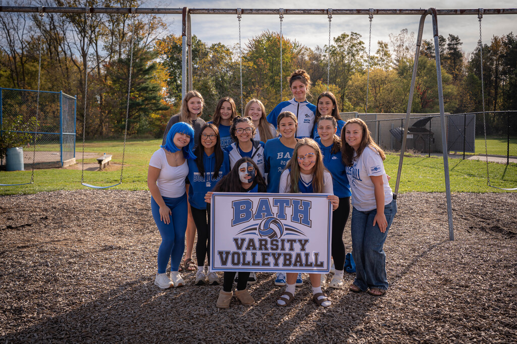 Girls  varsity volleyball team standing in front of 2 girls holding a sign that says Bath Varsity Volleyball. The are standing in front of a swing set on the playground.