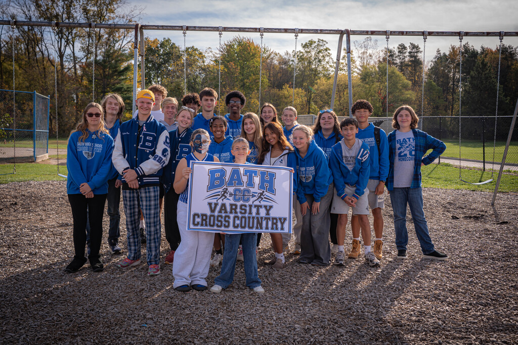 Varsity cross country team standing in front of 2 girls holding a sign that says Bath Varsity Cross Country.  The are standing in front of a swing set on the playground.