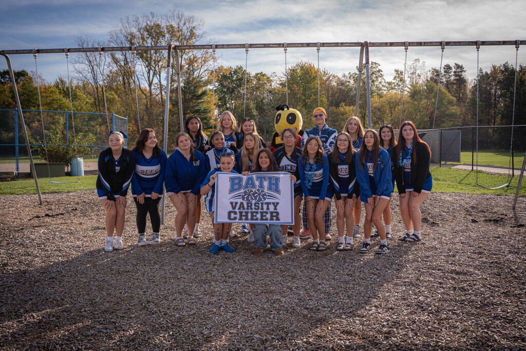 Girls varsity cheer team standing in front of 2 girls holding a sign that says Girls Varsity Cheer. The are standing in front of a swing set on the playground.