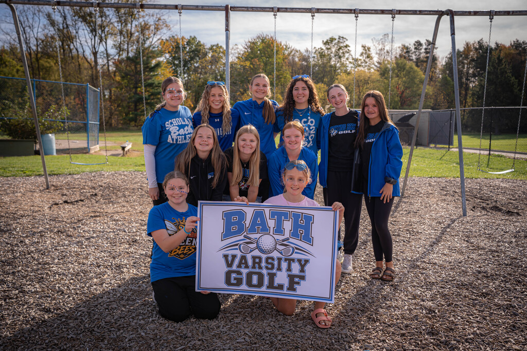 Girls varsity golf team standing in front of 2 girls holding a sign that says Bath Varsity Golf.  The are standing in front of a swing set on the playground.