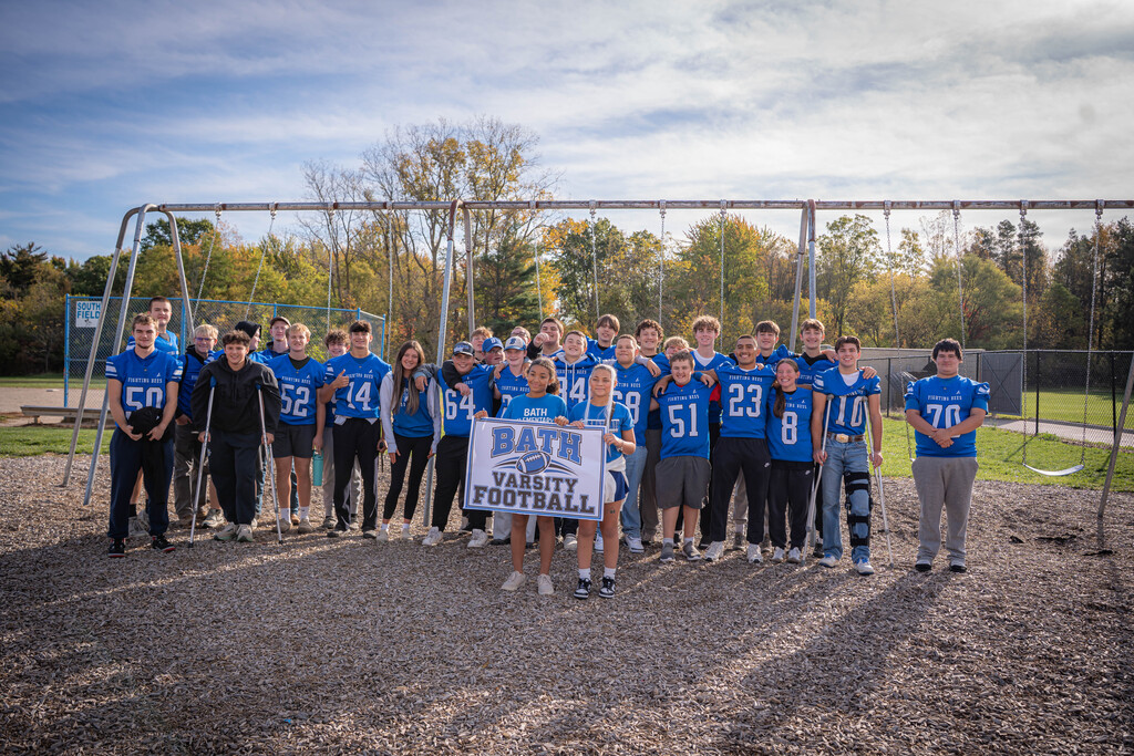 Boys varsity football team standing in front of 2 girls holding a sign that says Bath Varsity Football. The are standing in front of a swing set on the playground.