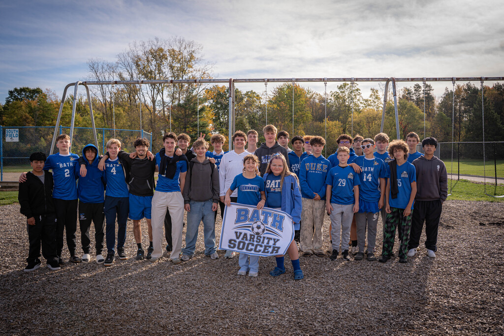 Boys soccer team standing in front of swing set on the playground. 2 girls standing in front of them holding a sign that says Bath Varsity Soccer