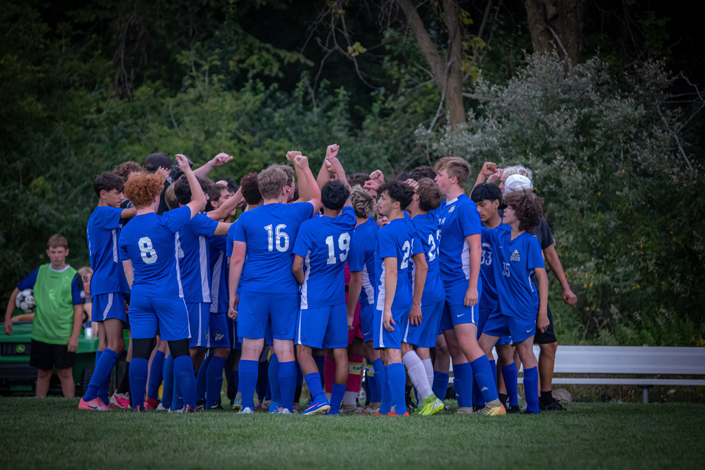 Boys soccer players wearing blue uniforms in huddle with arms raised