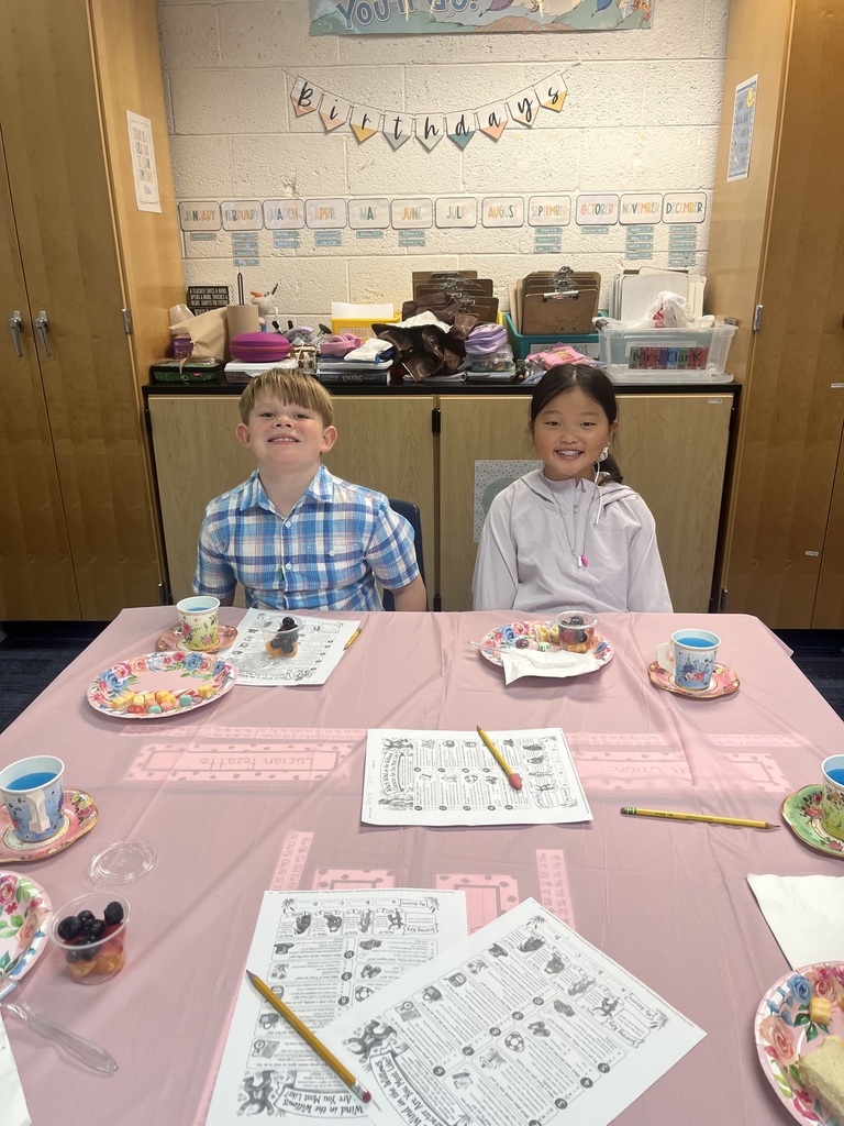 Girl and boy sitting at table with pink tablecloth that has papers, pencils and plates of food
