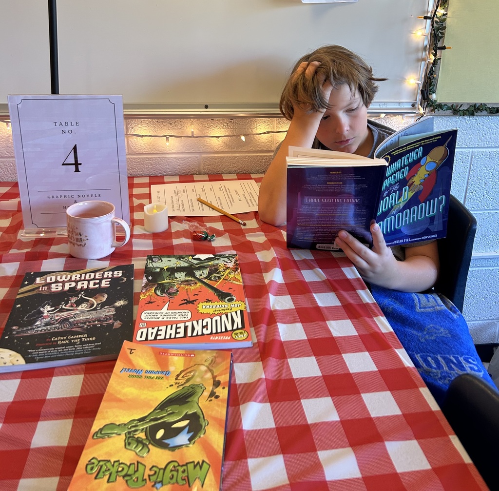 Boy sitting at table with red and white checkered table cloth reading a book. Several books are sitting on the table.
