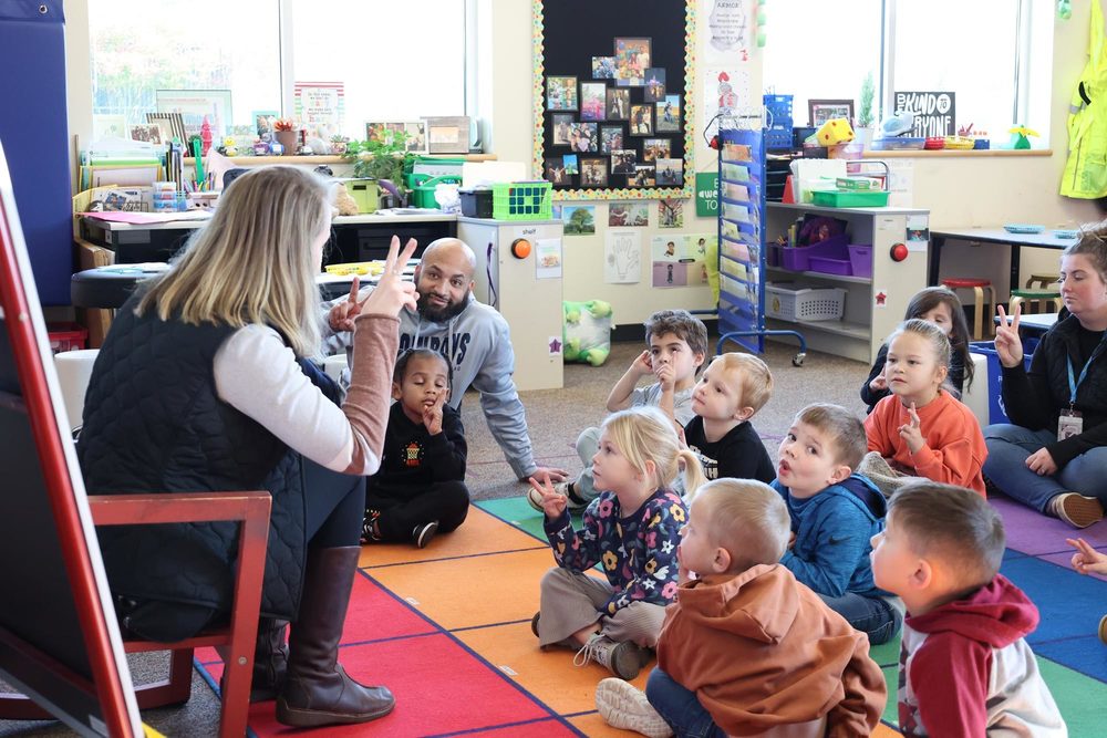 a tk teacher holds fingers up in the air while students are watching from  the carpet