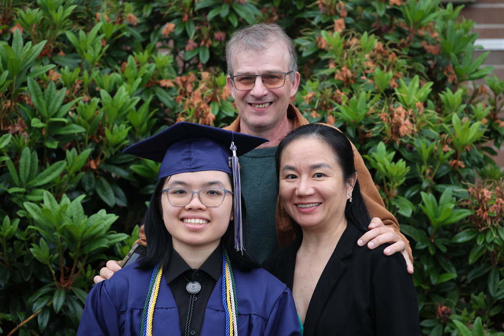 A young woman is standing with her mom and dad with greenery in the background