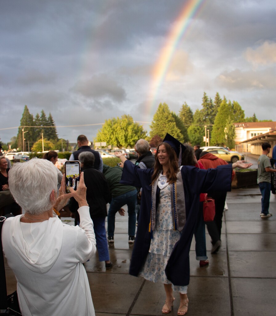 A rainbow and a happy arms wide open graduate smiles for her family member taking a photo of her