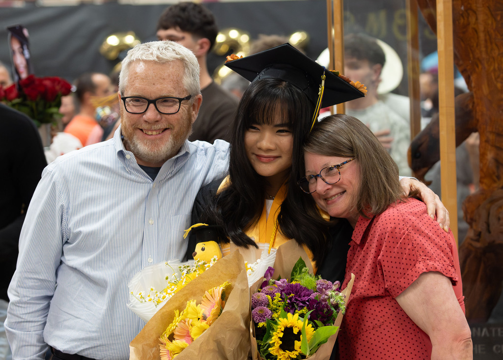 A young graduate smiles with her mom and dad while holding flowers