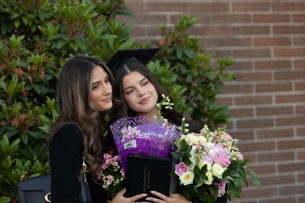 A young woman holds her flowers, graduation diploma, standing with her friend, both with beautiful smiles