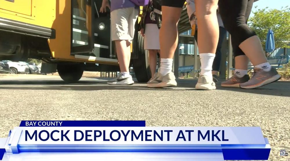 Students’ legs and feet are shown as they step onto a school bus, with a news caption reading “Mock Deployment at MKL” in Bay County.