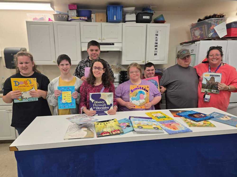 A group of students stand behind a table in a classroom, smiling and holding up children’s books, with more books displayed across the table in front of them.