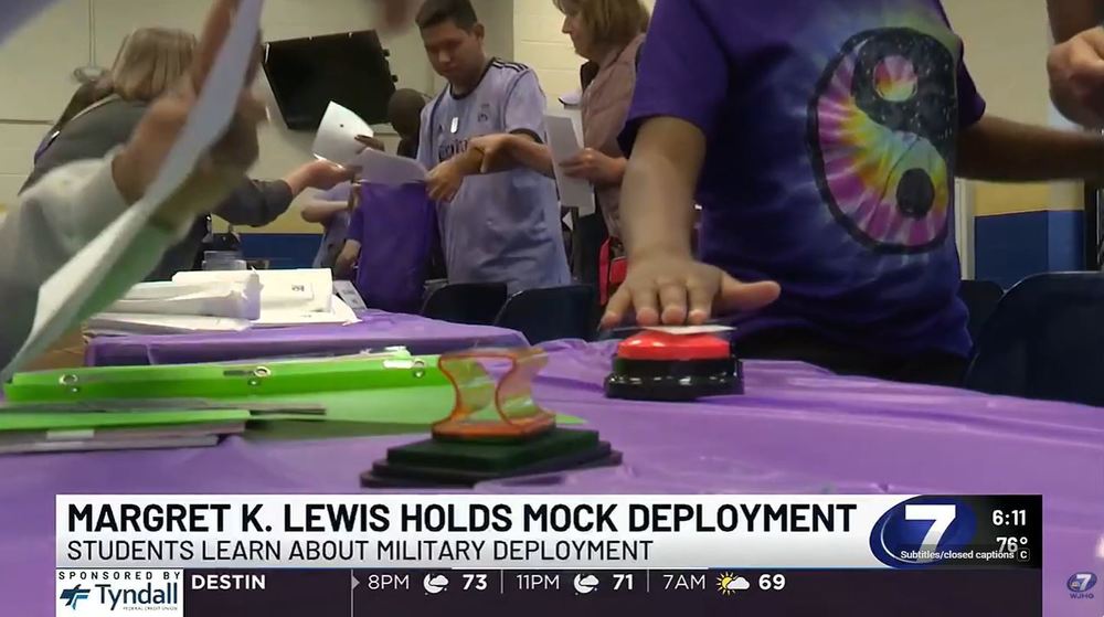 School news graphic showing emergency and supply kits—including bags, snacks, and medical items—displayed on a table during a mock deployment activity at Margaret K. Lewis School.