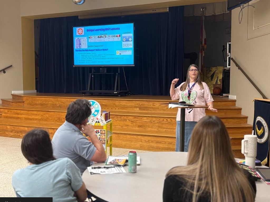 A staff member presents information on a screen about supporting student skills at home while families listen and take notes.