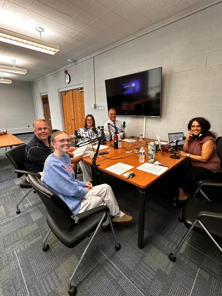Four adults and one student area seated around tables with microphones and notes as they prepare for a podcast recording.