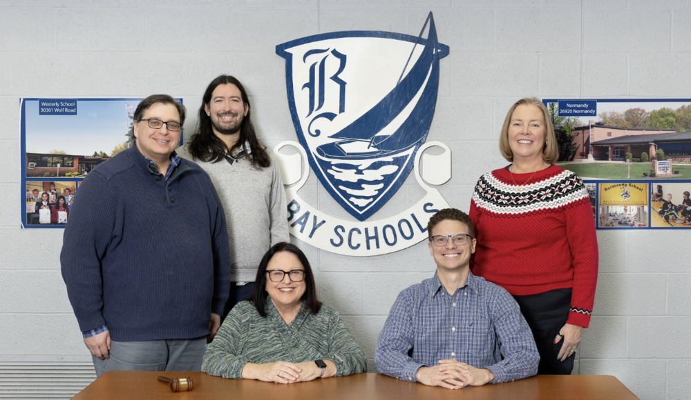 Board of Education Group Photo 2026. Five board members sitting and standing around a table with the Bay Village Schools emblem in the backround.
